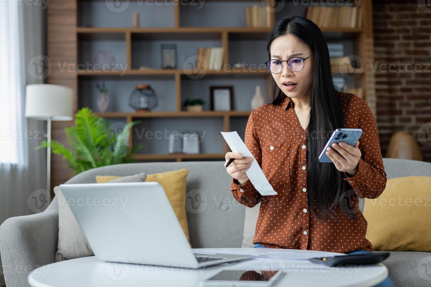 Asian woman at home surprised by high bills, holding smartphone and looking at laptop. Illustrates financial stress and technology use for budgeting and managing expenses. photo