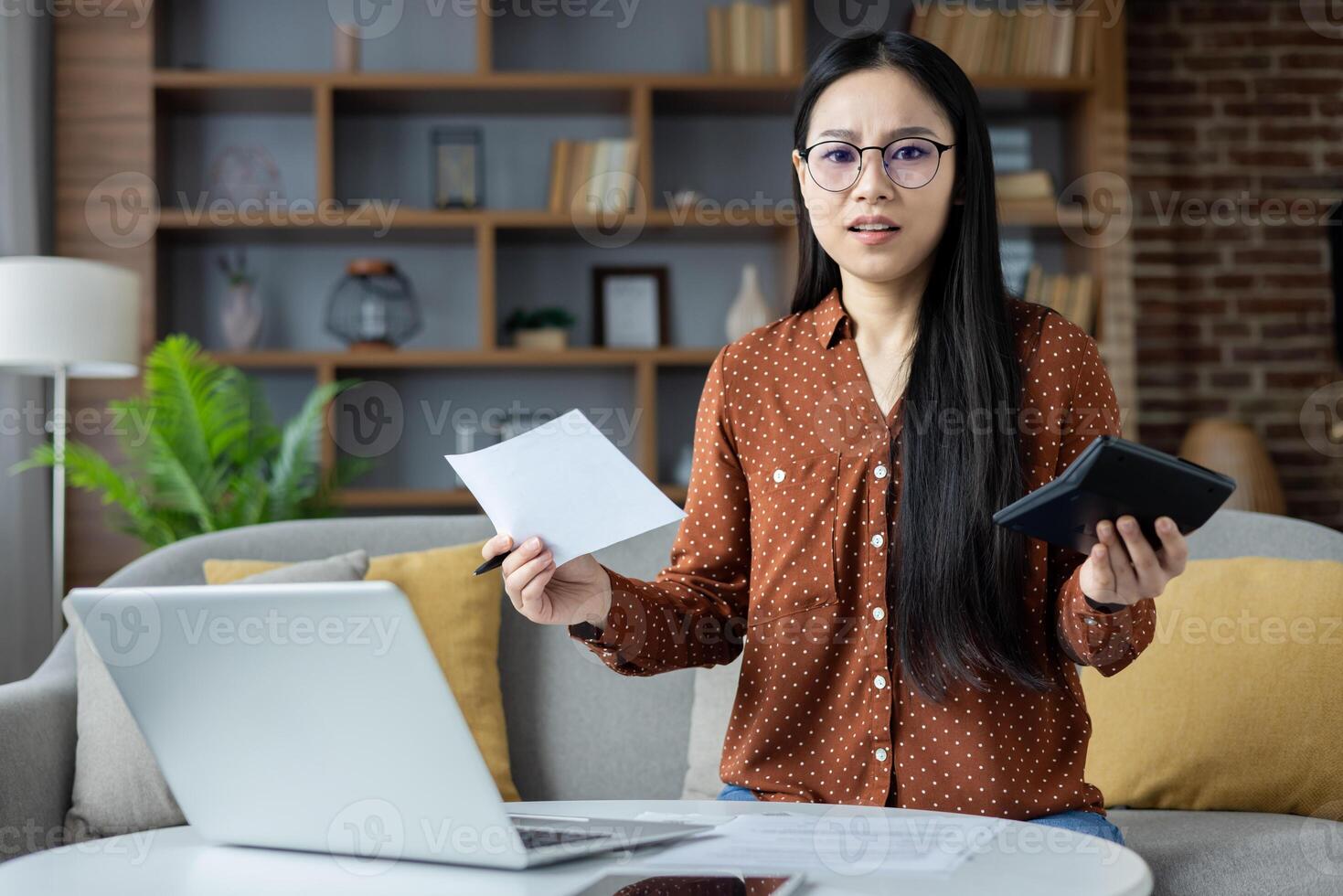 Asian woman at home looks puzzled over finances, holding paper and calculator. Laptop on table representing remote work challenges and financial planning stress. photo