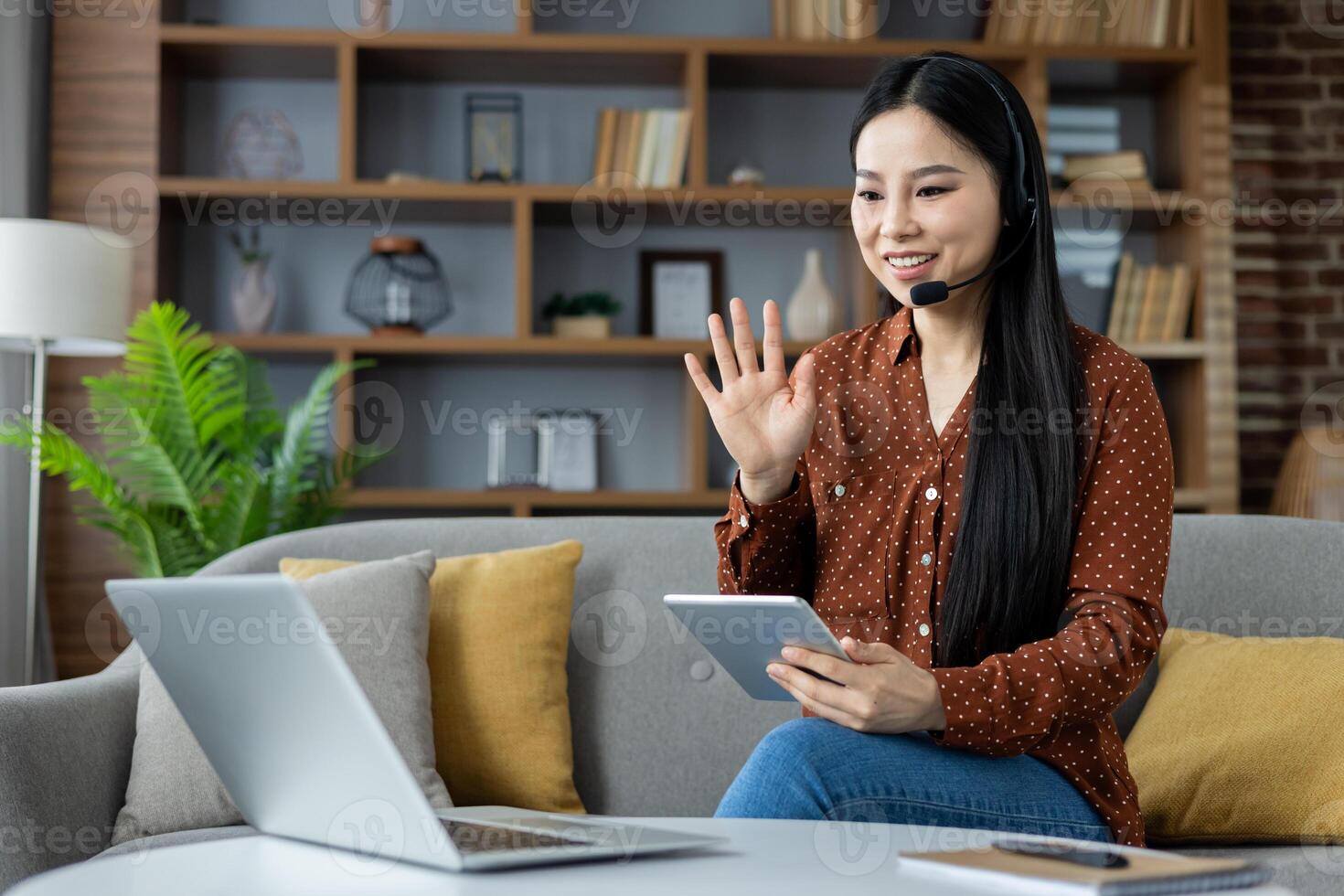 Asian woman using laptop and tablet for call at home, wearing headset, smiling and waving during online meeting. Cozy living room setting enhances a work-from-home atmosphere. photo