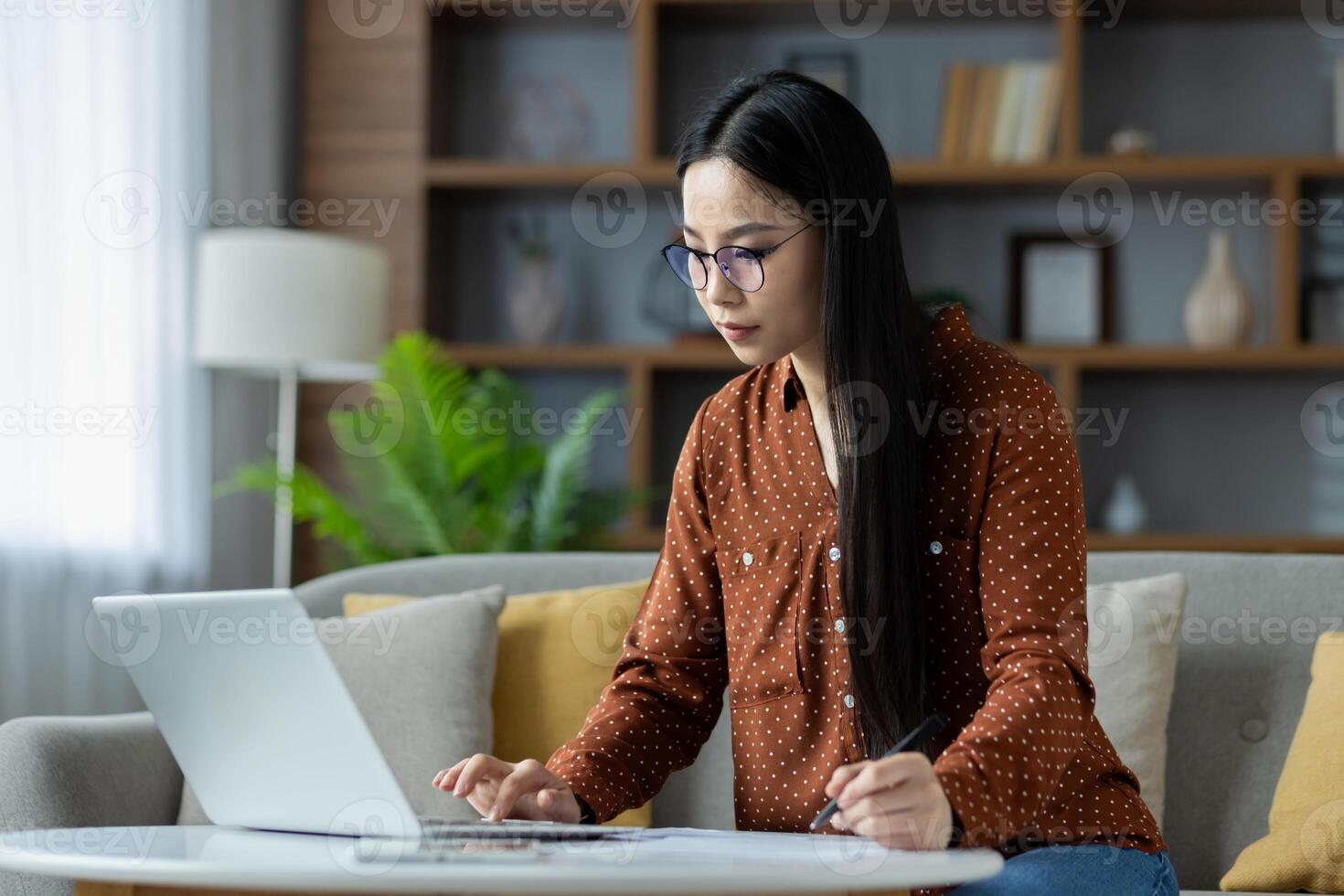 Asian woman working from home on laptop, deeply focused in stylish living room. She uses pen and paper for notes, showcasing remote work lifestyle and concentration in cozy environment. photo