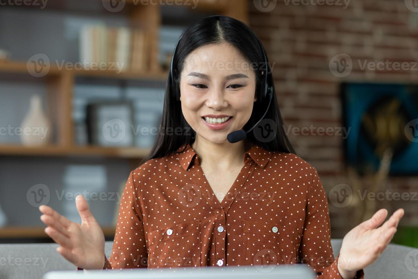 Asian woman in a virtual meeting, wearing headset, smiling and gesturing. Shows remote work and online communication in comfortable home setting. Captures professional yet relaxed atmosphere. photo