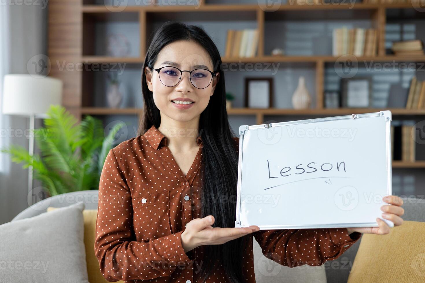 Confident Asian woman holding whiteboard with 'Lesson' text, ready for online teaching or virtual training session in comfortable home setting. Wearing eyeglasses and professional attire. photo