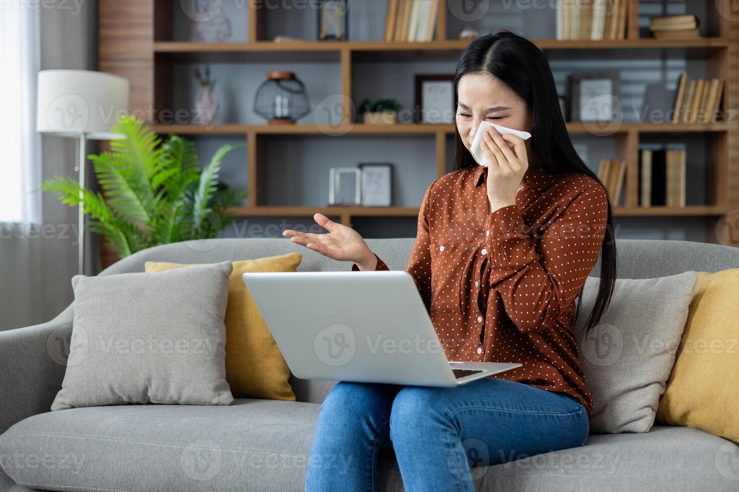 Asian woman using laptop on sofa, holding tissue, appears emotional, possibly chatting. Wearing casual attire, setting suggests home environment with emphasis on comfort and technology. photo