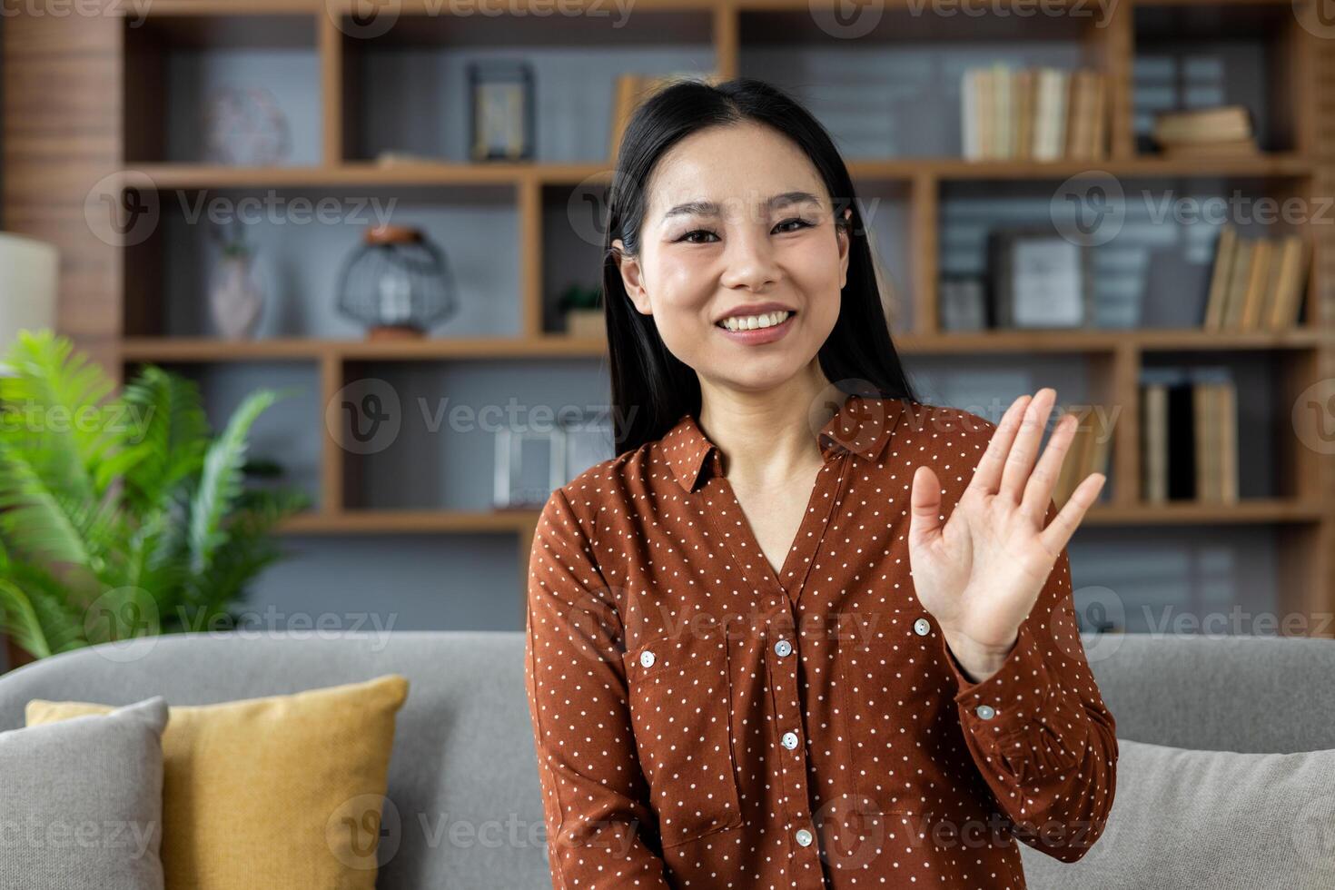 Asian woman sitting on sofa waving warmly at camera. She's wearing a brown polka dot shirt, expressing friendliness and happiness. Ideal for virtual meetings, online communication, home setting. photo
