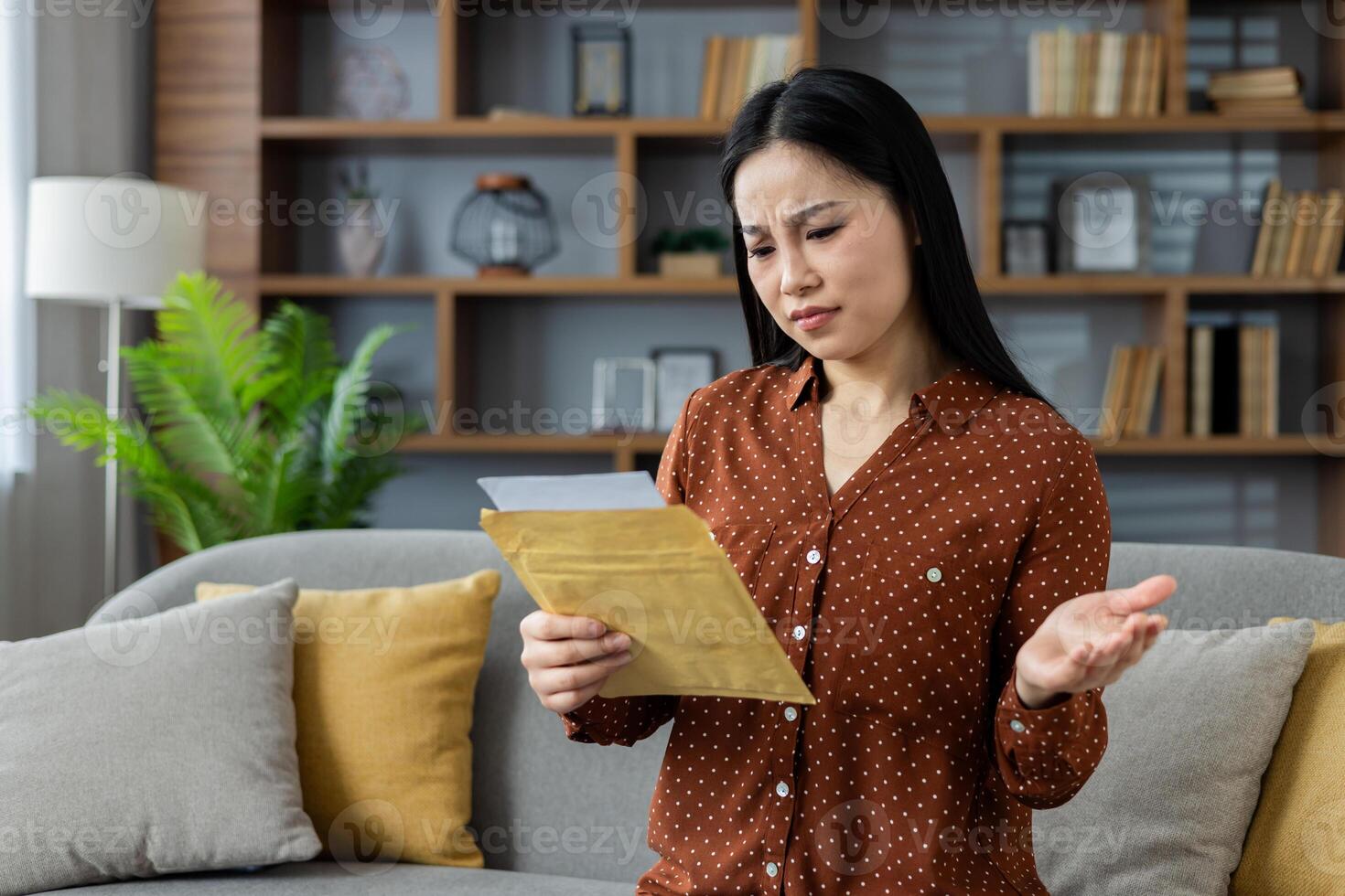 Asian woman at home reading a letter with confused expression on sofa. She appears thoughtful and concerned, reacting to unexpected news in a modern living room setting. photo