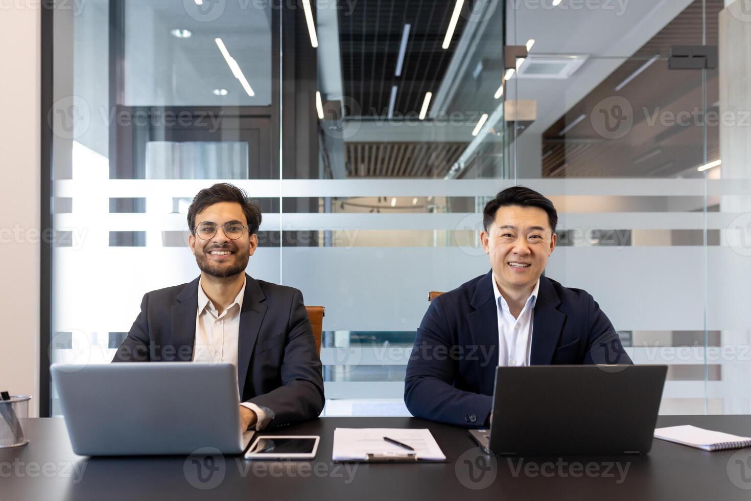 Two professional men sitting at a desk in a contemporary office, smiling confidently. They are working with laptops and have documents and a smartphone in front of them photo
