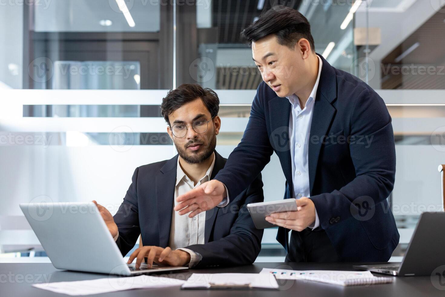 Two business professionals engage in a collaborative discussion in a modern office environment. One stands holding a tablet, while the other works on a laptop. The setting highlights teamwork. photo