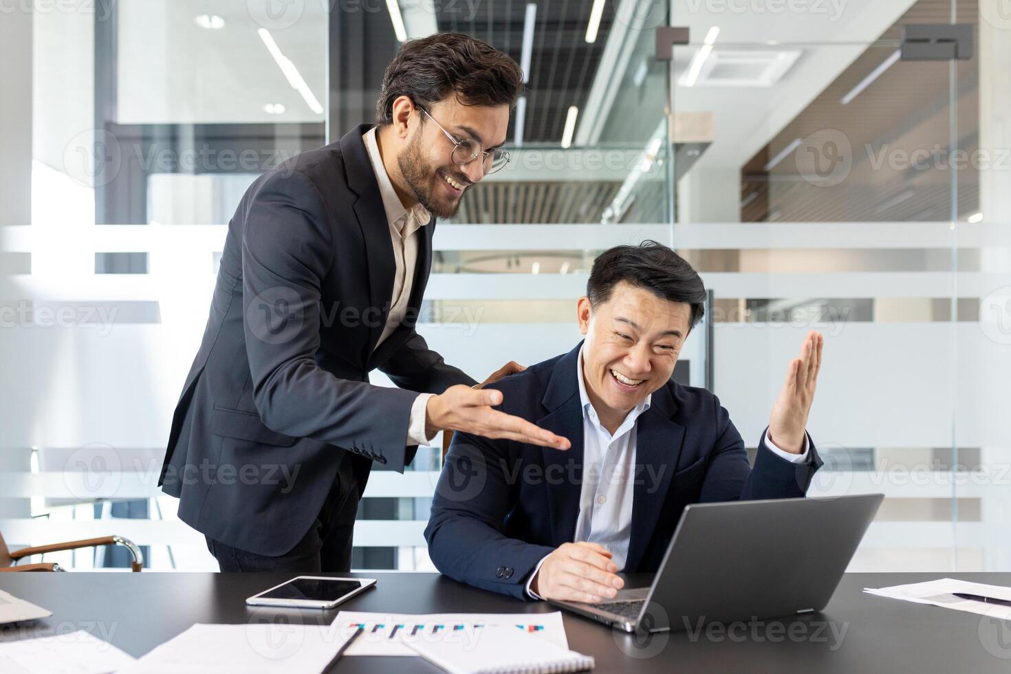 Two businessmen are joyfully celebrating a success in a modern office setting. One is using a laptop, while the other gestures excitedly. Their expressions show happiness and teamwork. photo