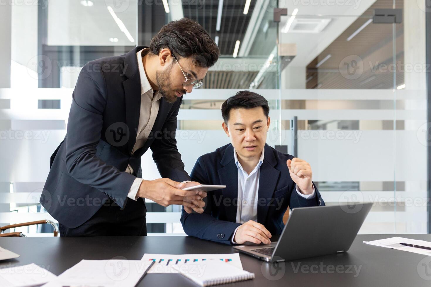 Two professionals collaborate in modern office using laptop and smartphone, emphasizing teamwork, technology, productivity. Engaged managers discussing ideas, showcasing business strategy photo