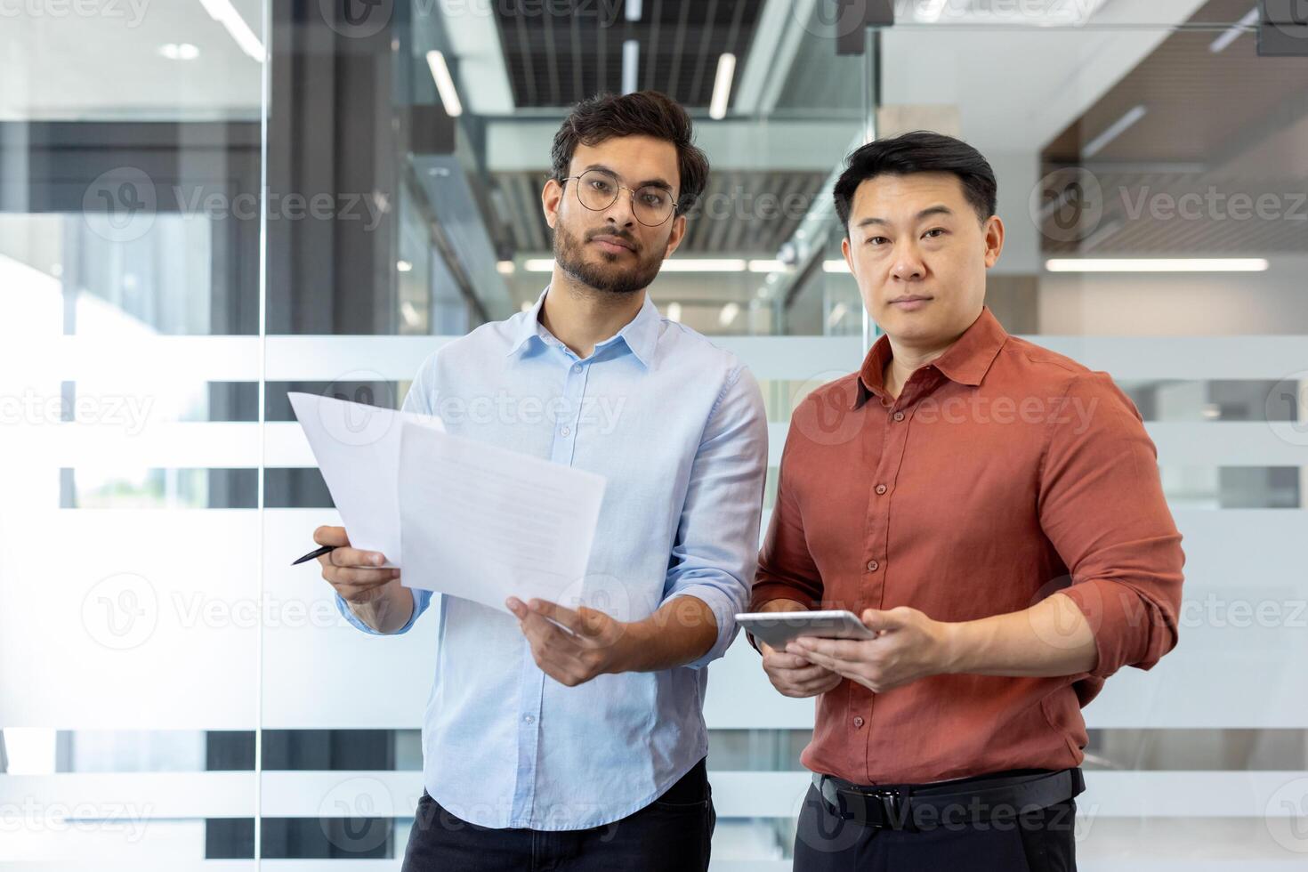 Two businessmen stand in a modern office holding documents and a tablet. They engage in a business discussion, focused on strategy and collaboration. T photo