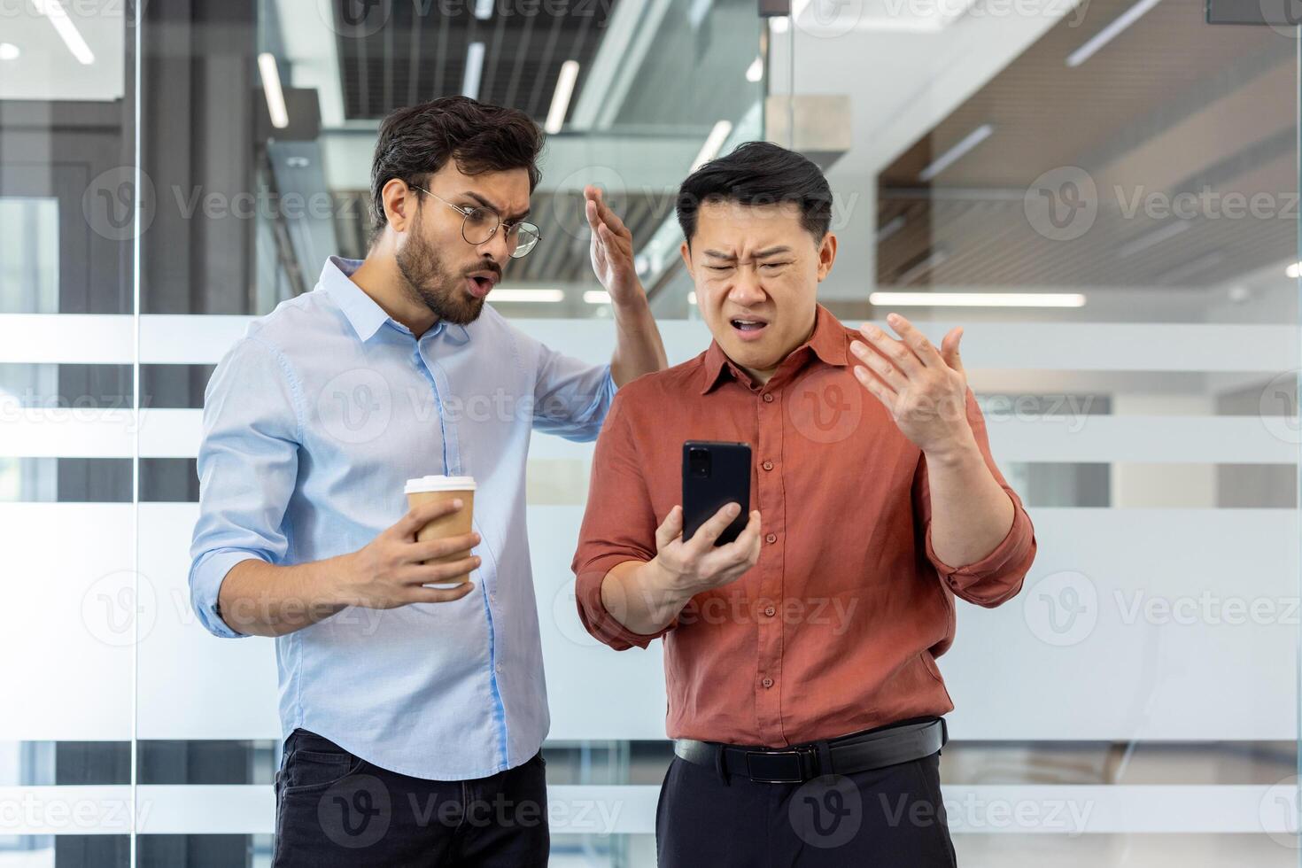 Two business professionals express frustration over unexpected phone problem in modern office setting. One holds coffee cup, both focus on smartphone displaying issue, conveying stress and urgency. photo