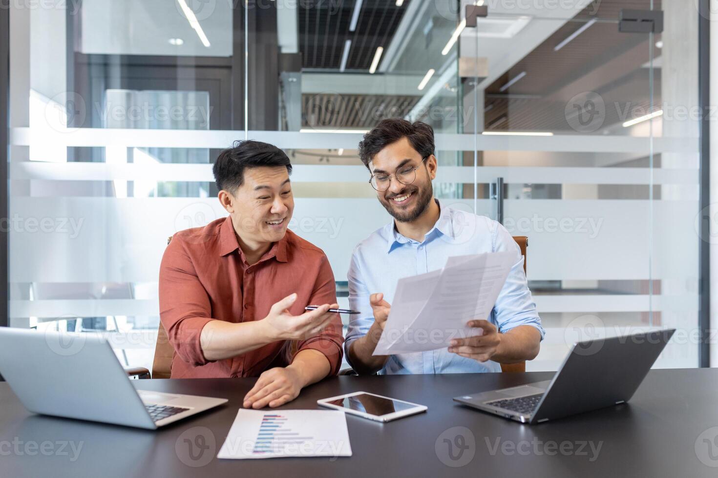 Two colleagues are sitting in a contemporary office discussing paperwork. They are using laptops and a tablet, reflecting a productive and collaborative work environment. photo