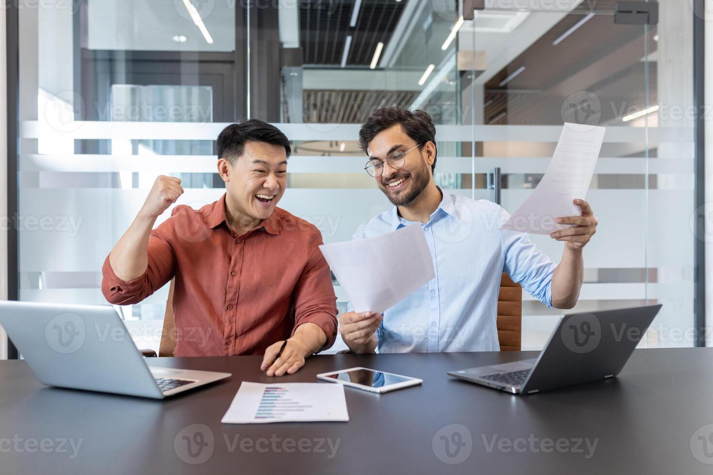 Business colleagues celebrate success with laptops and documents. Smiling coworkers review project results on paper and phone, highlighting teamwork and achievement in modern office. photo