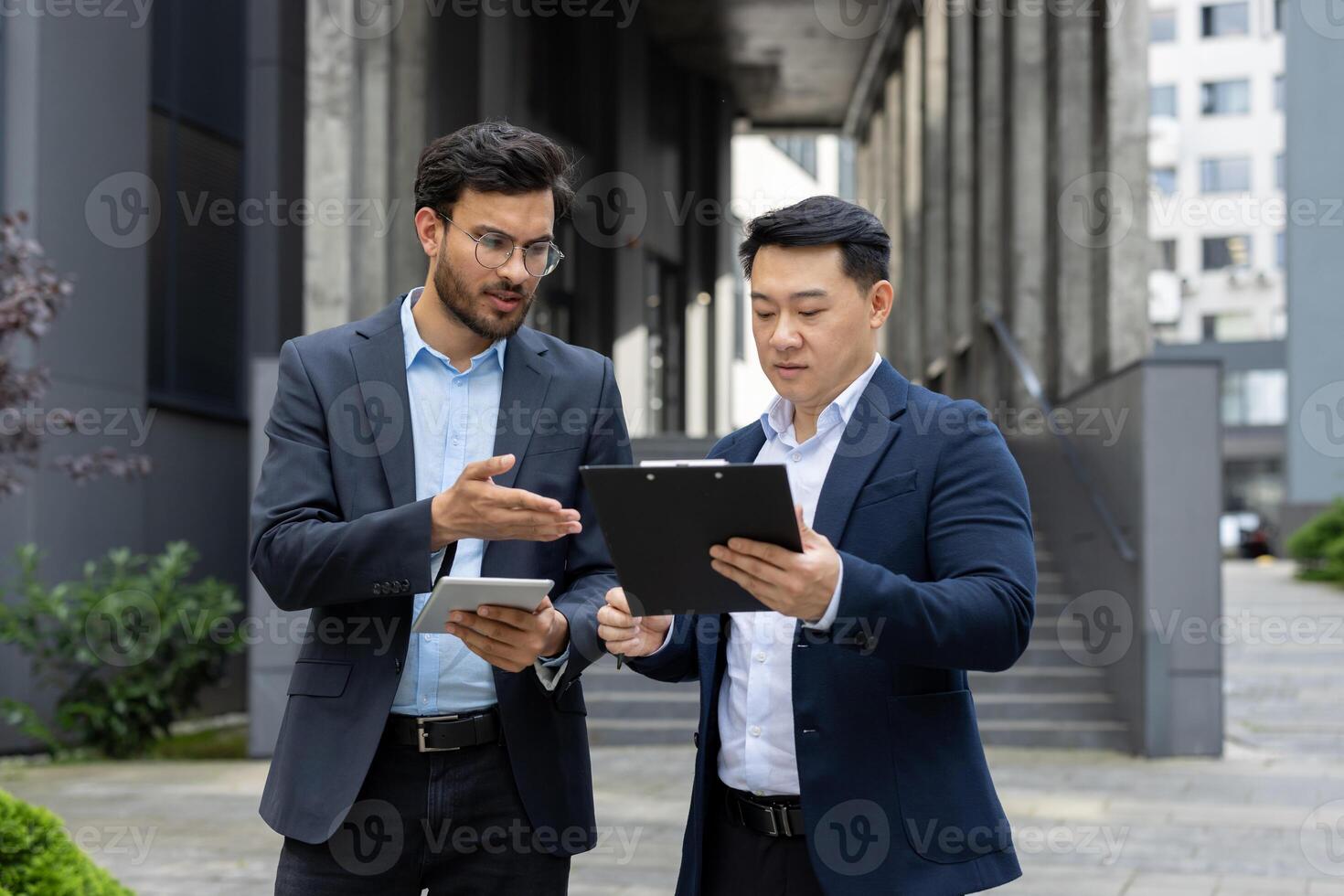 Businessmen discussing work in corporate environment. One holding tablet, another with clipboard, focused on conversation. Modern setting enhances business strategy context. photo
