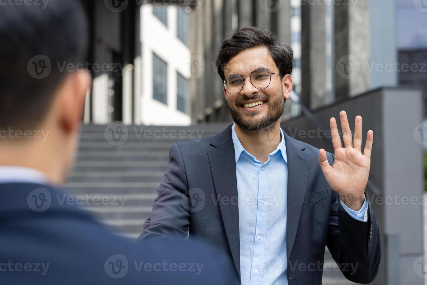 Businessman in suit greeting colleague with friendly smile and wave. Outdoor meeting in city, showcasing professional interaction and positive connection during workday. photo