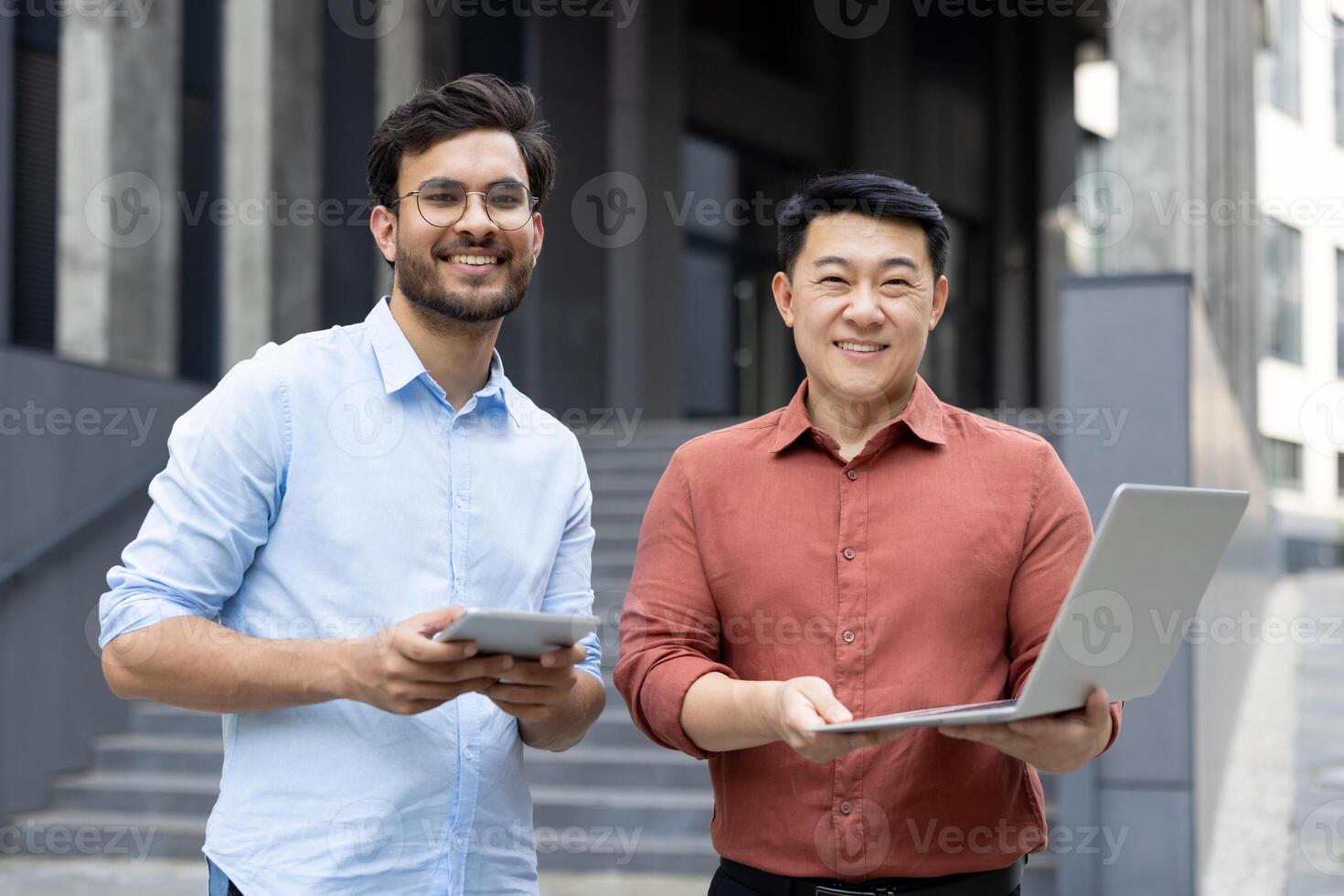 Two professionals, equipped with a tablet and laptop, are smiling while working outdoors. This scene suggests a collaborative business environment with a focus on technology and teamwork. photo