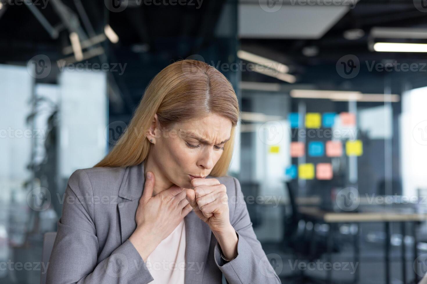 Mature woman businesswoman coughing in office, exhibiting signs of discomfort or health issue. Expression indicates potential stress or illness amidst professional setting. photo
