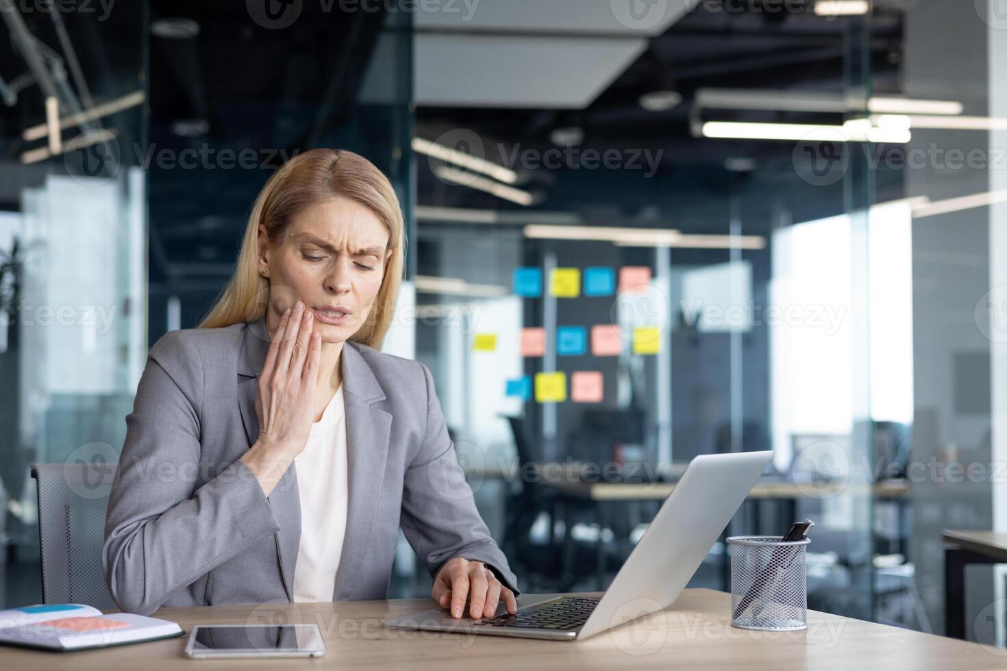 A mature businesswoman, feeling discomfort, touches her face while working on a laptop. She appears to be experiencing tooth pain in a modern office setting. Her expression suggests discomfort photo