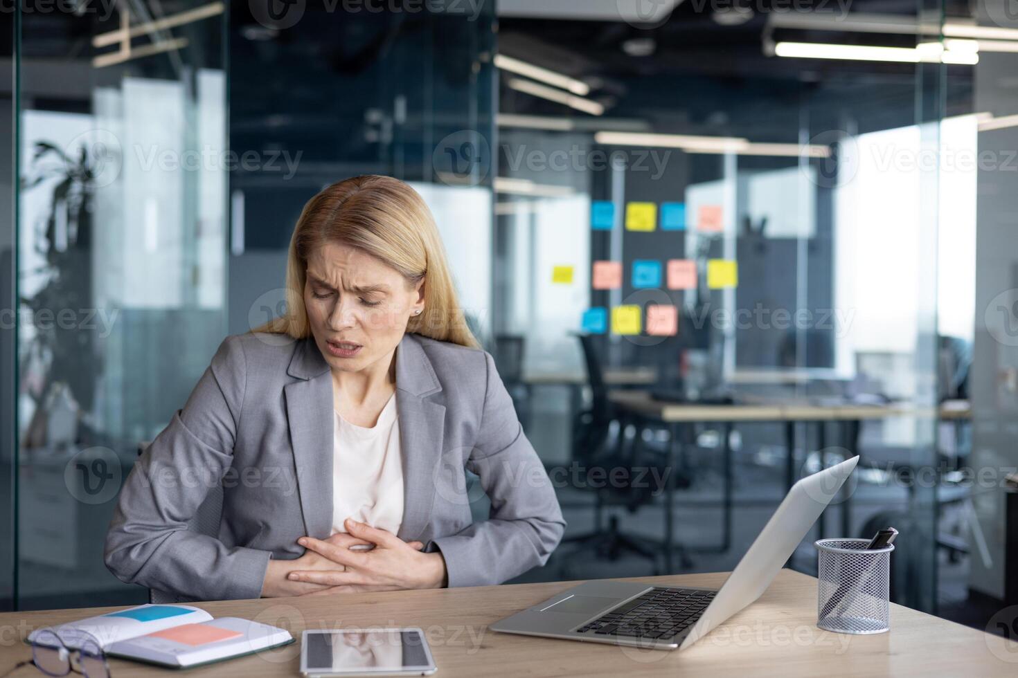 A mature woman businesswoman sits at a desk in a modern office, experiencing discomfort. She appears concerned, with her hand on her stomach. The setting includes a laptop, documents, and a phone. photo