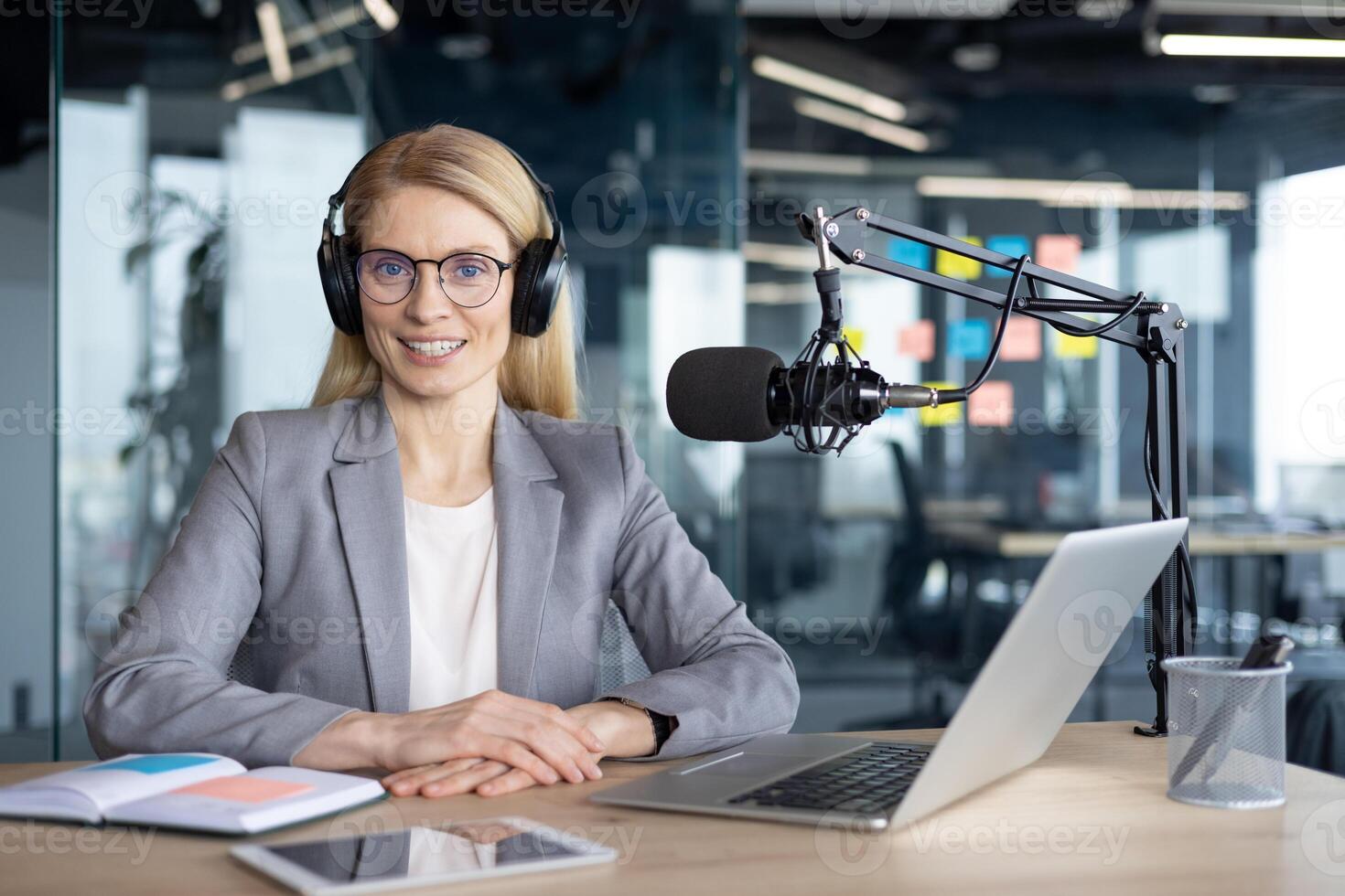 Mature businesswoman using microphone for podcast recording in modern office. Headphones and laptop on desk suggest professional setup for broadcasting or virtual communication. photo