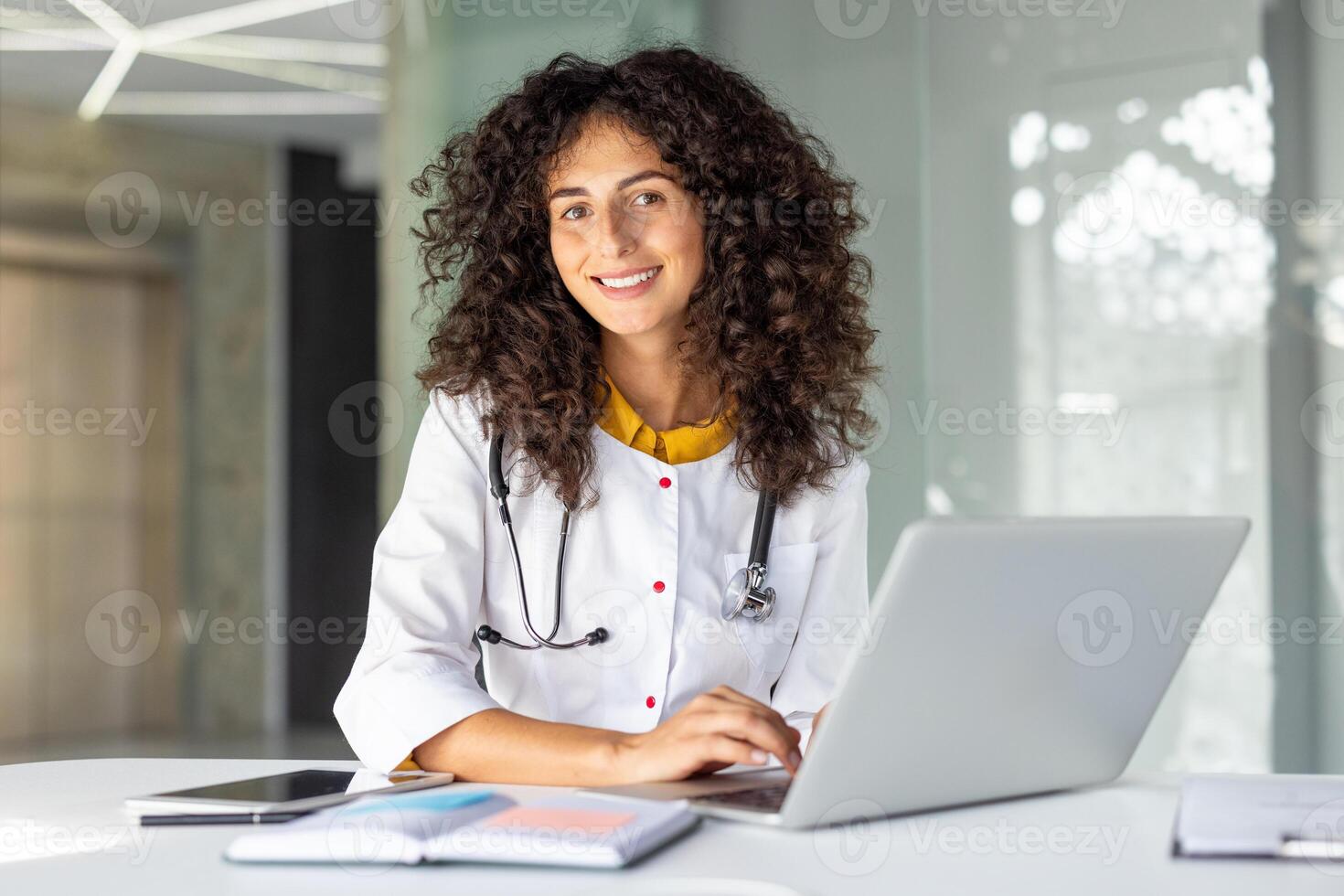 A confident female doctor with a stethoscope uses a laptop in a modern office. She is smiling, wearing a white coat, and appears professional. The setting suggests healthcare, technology photo