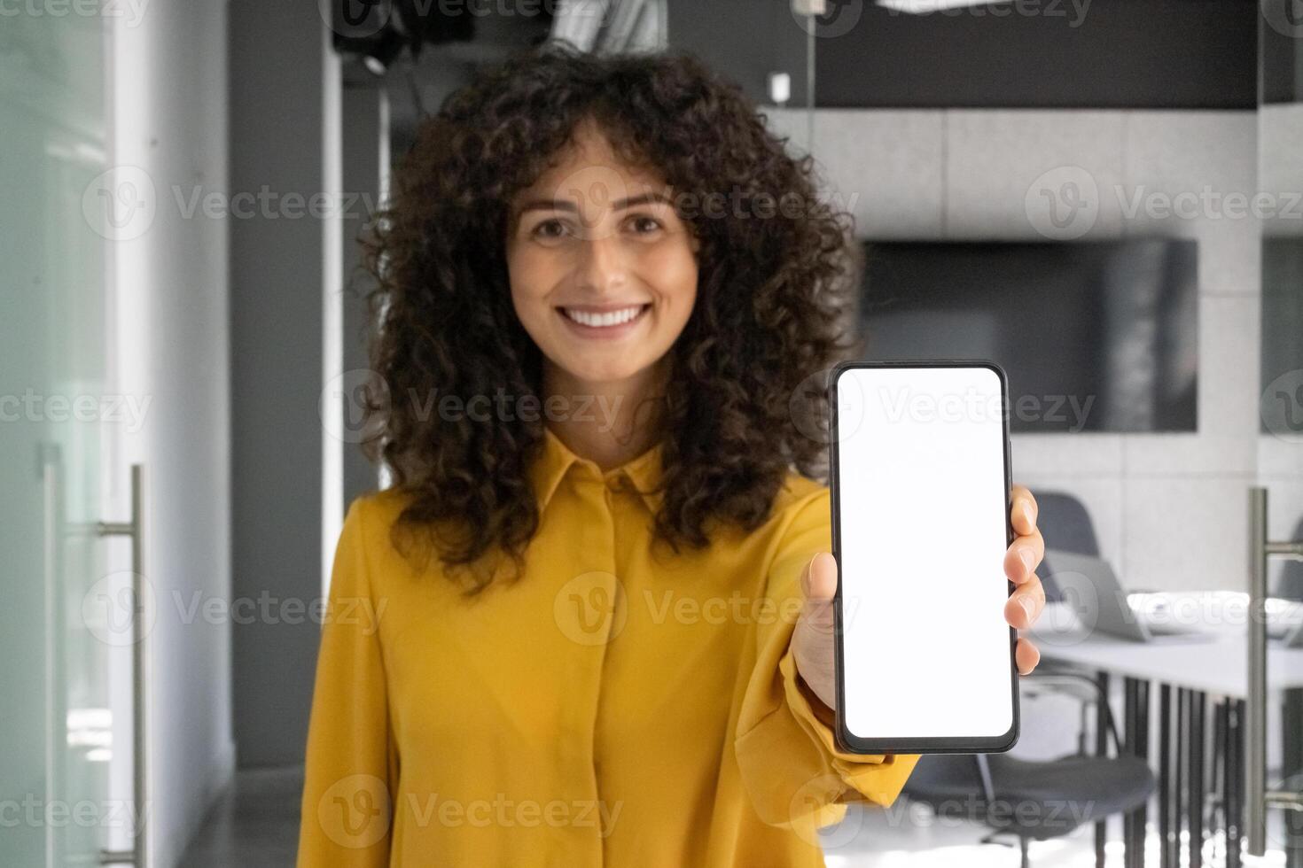 Latin American businesswoman in office holding smartphone with blank screen, smiling confidently. Concept of technology use, app promotion, and professional communication in workplace with phone. photo
