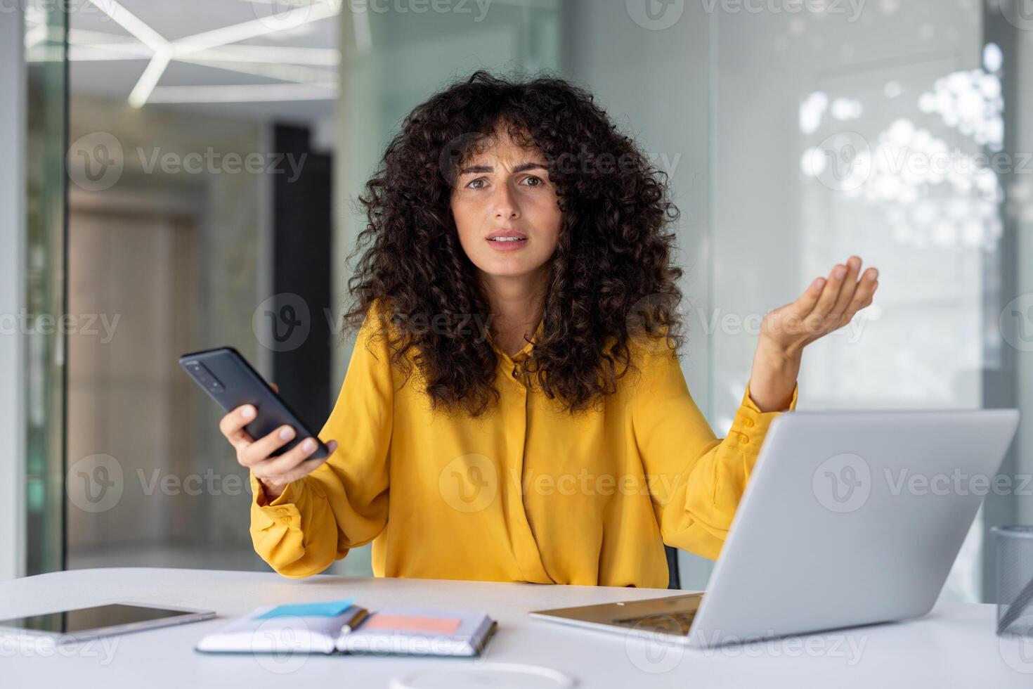 Curly-haired woman in bright office experiences frustration, holding smartphone and gesturing in confusion. Laptop on desk suggests work stress or technology issue. Emotions visible on face. photo