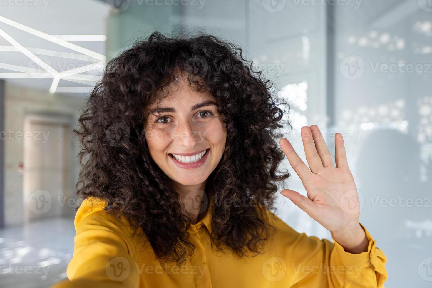 Woman with curly hair waves and smiles in office, capturing selfie with phone. Energy and friendliness radiate as she wears yellow shirt, set in modern business environment. photo