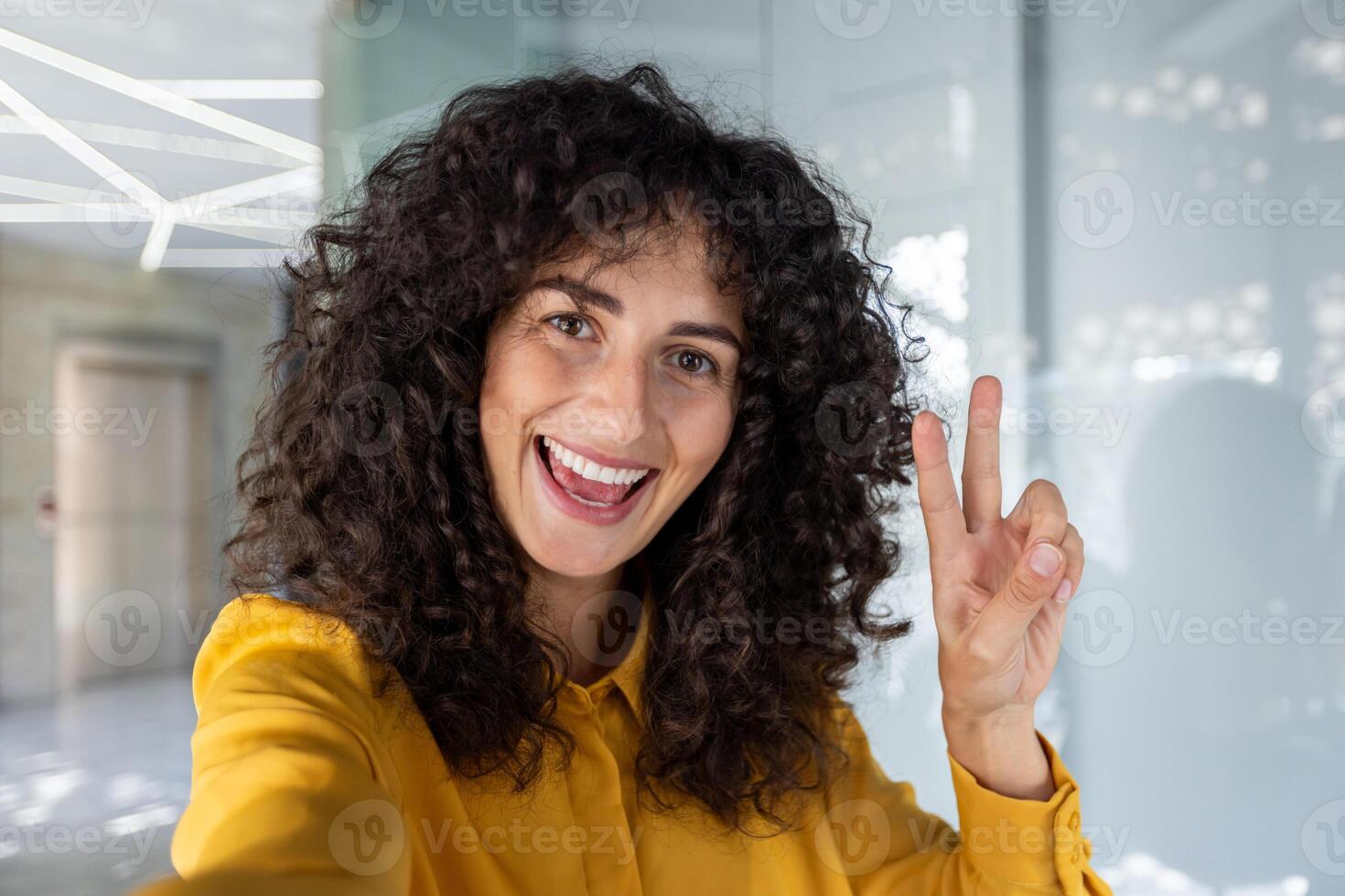 Woman takes selfie indoors. Peace sign and yellow shirt add to cheerful, positive mood. Capturing joyful expression using phone, promoting happiness in modern lifestyle setting. photo