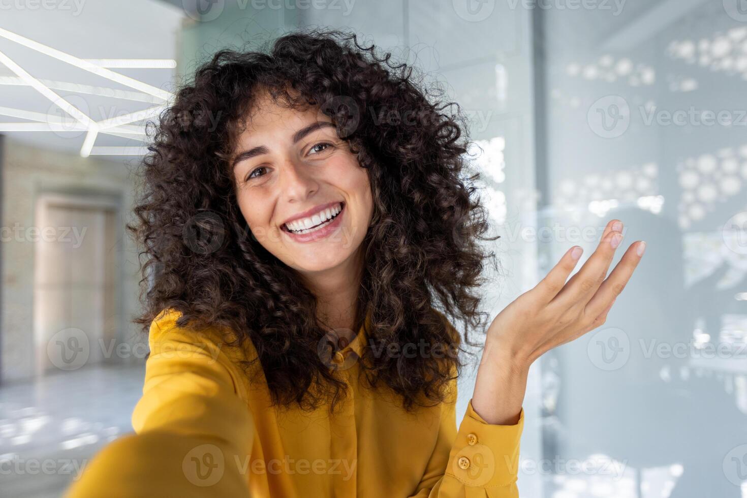 Smiling woman with curly hair taking selfie in bright yellow blouse. Her cheerful expression conveys positivity, while office atmosphere suggests a professional, welcoming vibe. Phone in hand. photo