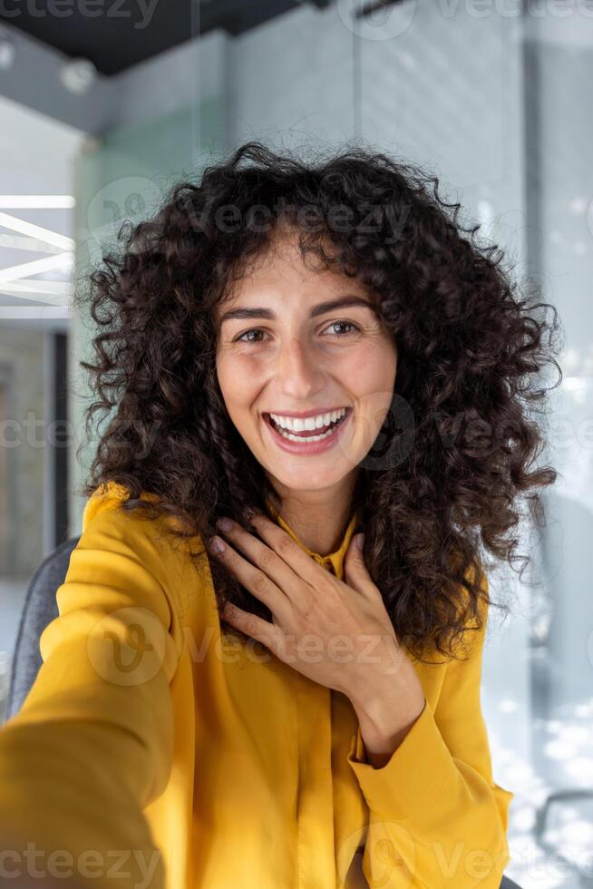 A cheerful woman takes a smartphone selfie in an open-office environment. Her friendly smile and outgoing energy are perfect for illustrating concepts like happiness and connection. photo