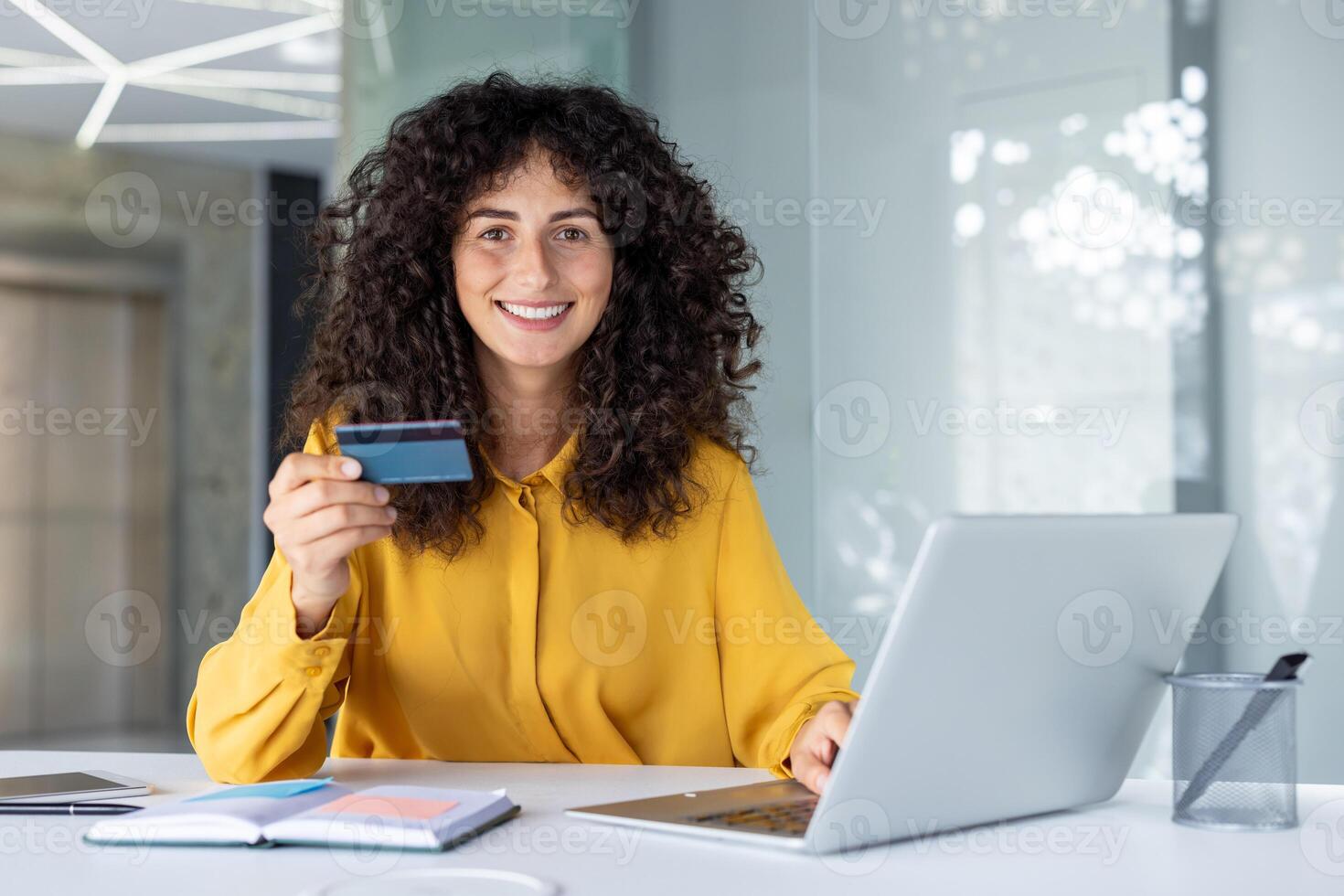 Smiling woman in yellow blouse holding credit card at office desk, using laptop for online shopping or banking. Concepts of business, finance, tech, and digital transactions. photo