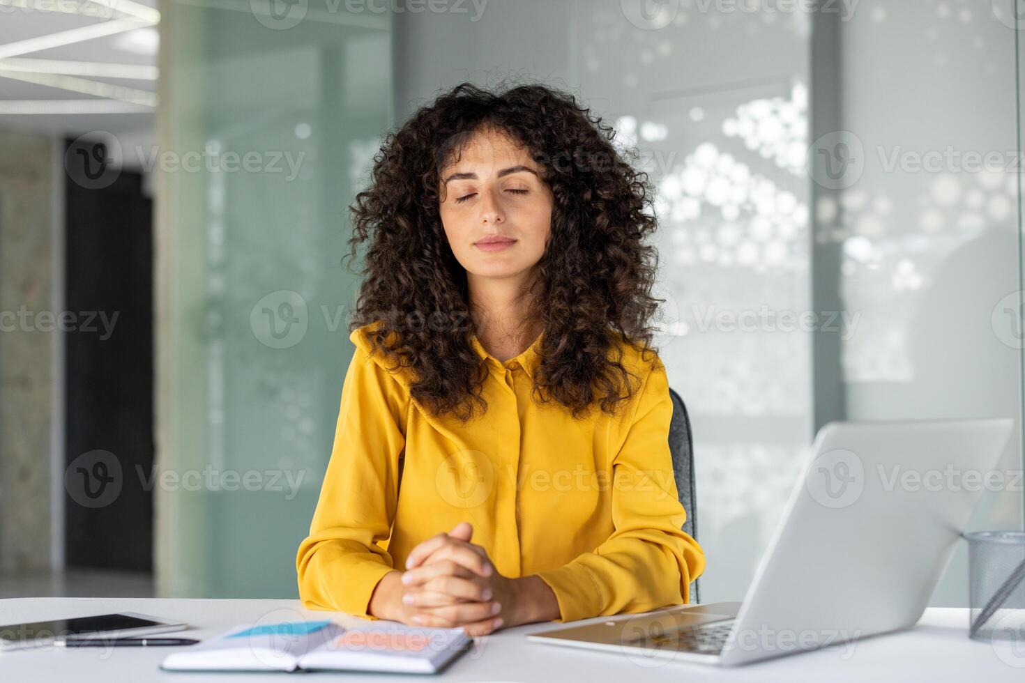 Businesswoman embracing tranquility at a desk, eyes closed, practicing meditation for mindfulness and stress relief. Laptop and notepad symbolize modern office setting and work-life balance. photo