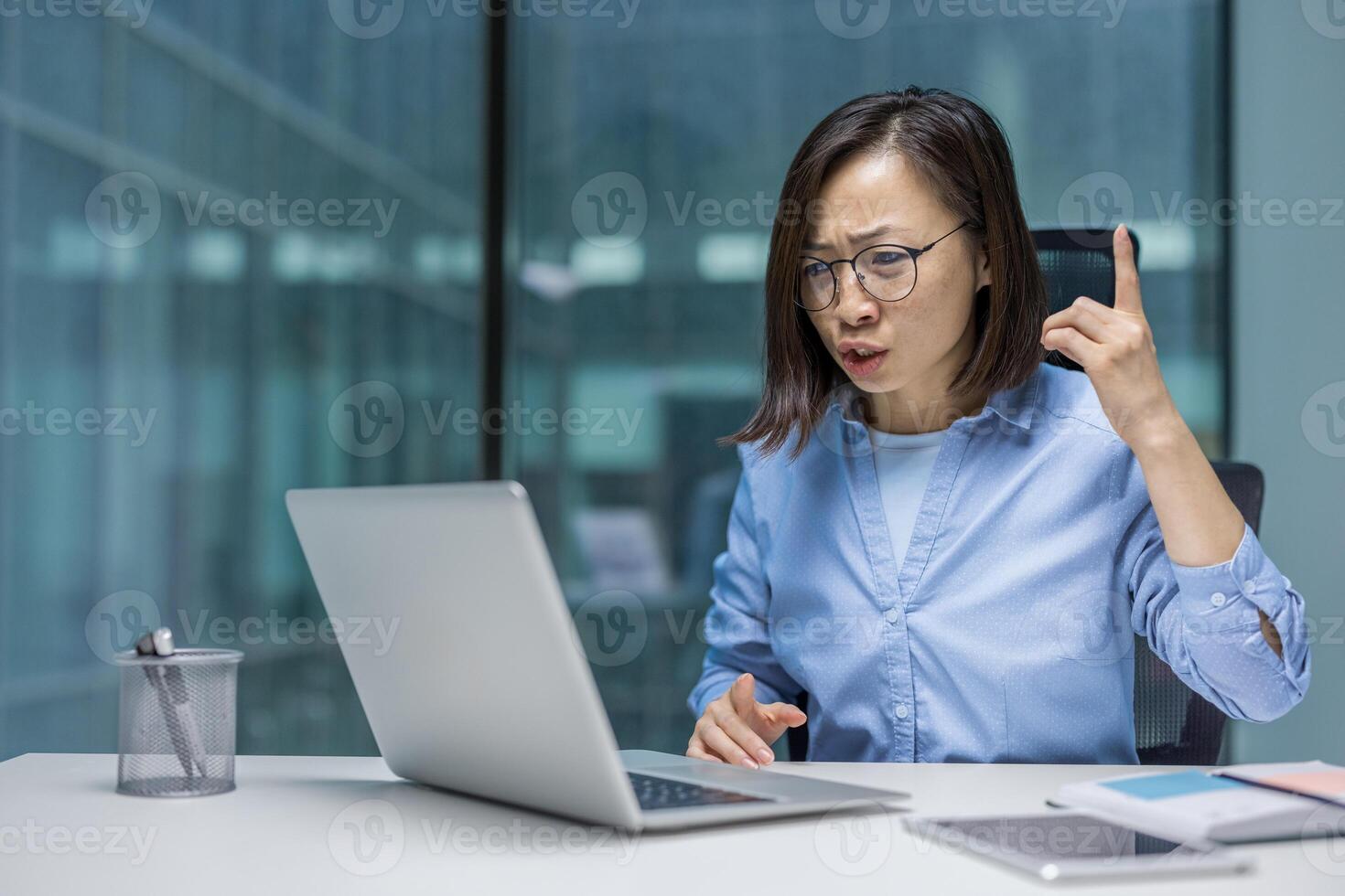 Asian woman at office desk expressing frustration looking at her laptop. Her suspicious expression and finger gesture emphasize her annoyance, perhaps due to a challenging task or communication. photo