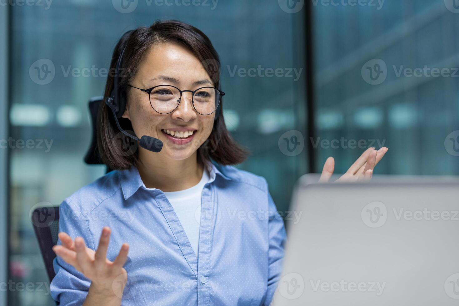 Asian woman wearing a headset is actively participating in a virtual meeting. She is gesturing with her hands and smiling, reflecting communication and engagement in a professional environment. photo