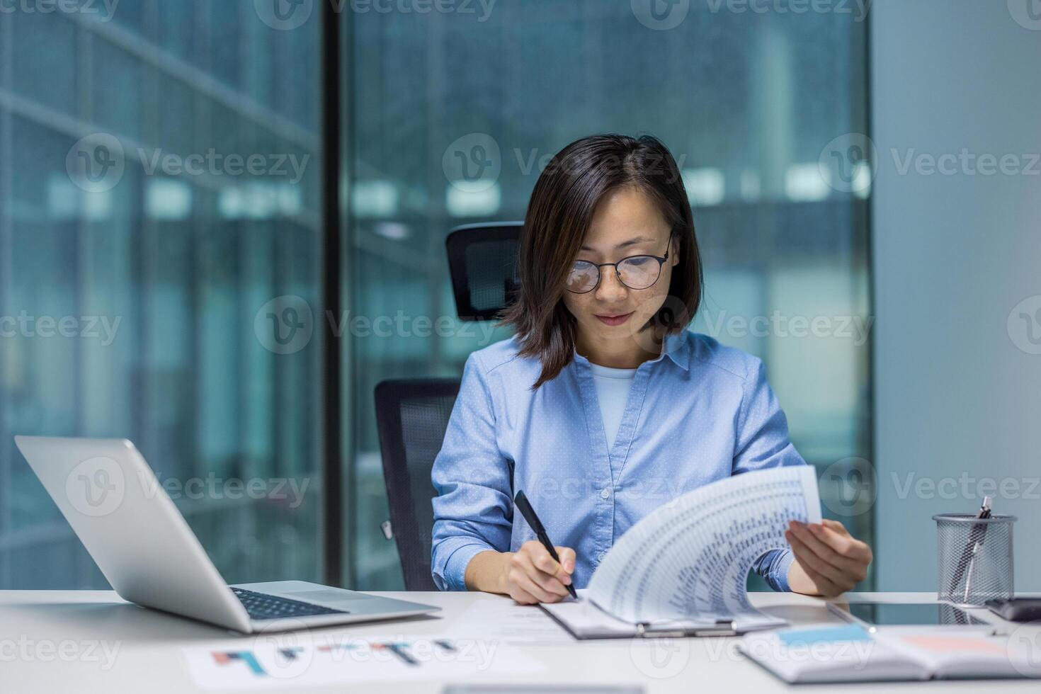 Asian woman concentrates on analyzing data at her office desk. She reviews documents while using a laptop, exemplifying professionalism and focus in a modern workspace. photo