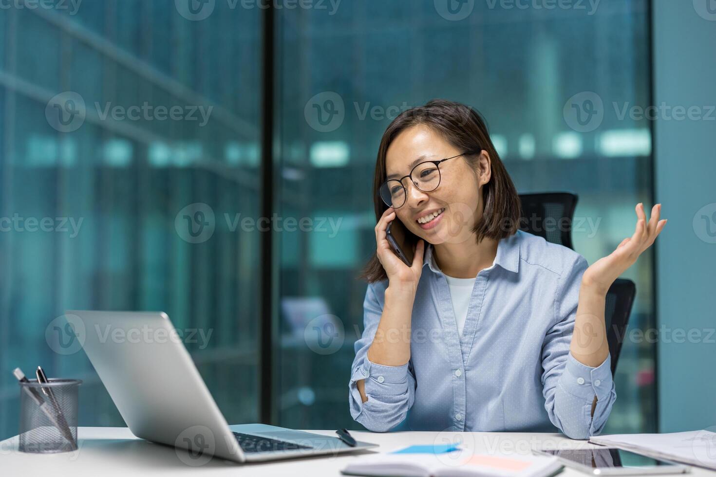 Asian businesswoman on phone call in office. Smiling and confident while using laptop, showcasing modern work environment and communication. Captures professionalism and productivity. photo