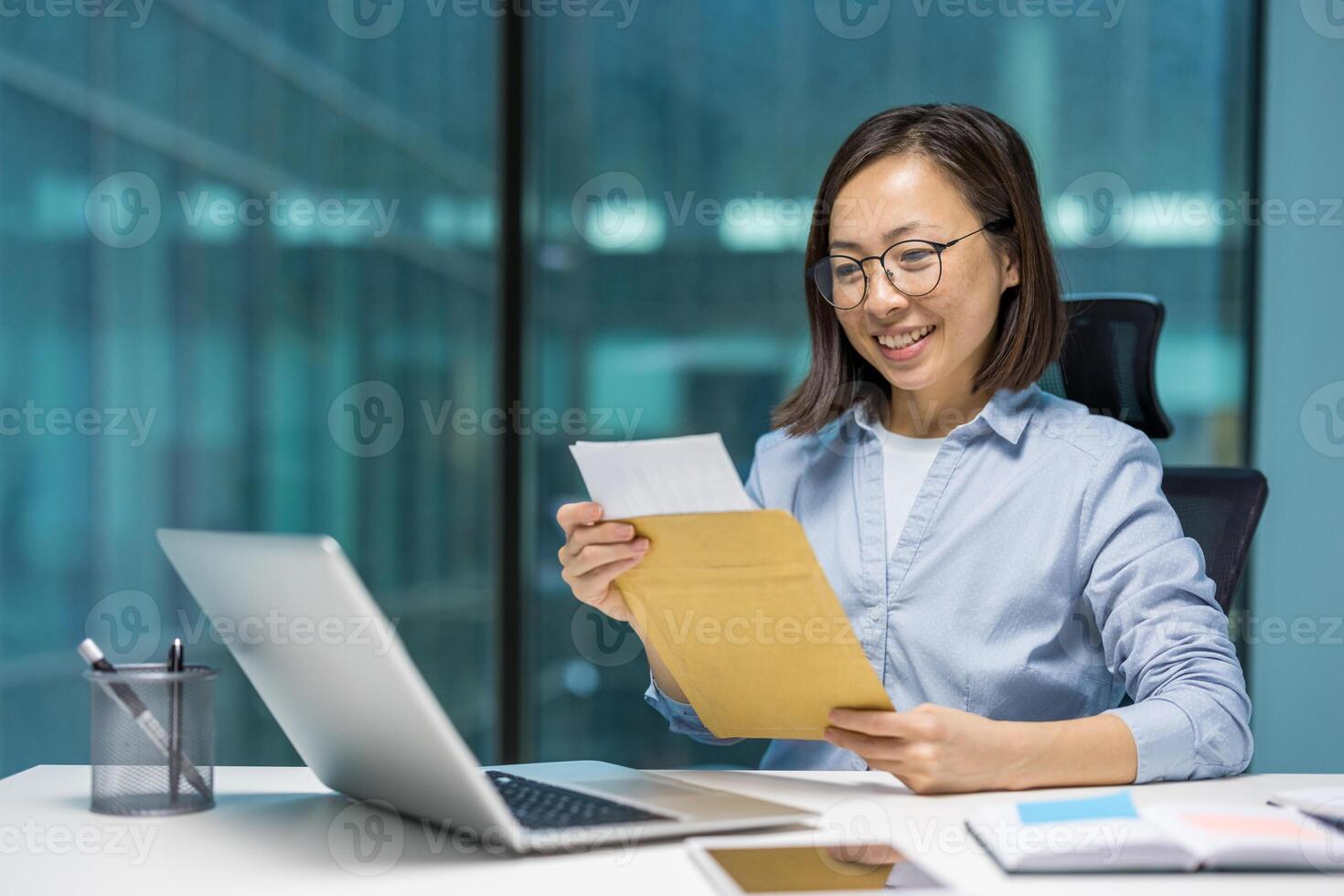 Asian businesswoman reads good news from envelope at office desk, surrounded by laptop and documents, showcasing professional success and positivity photo