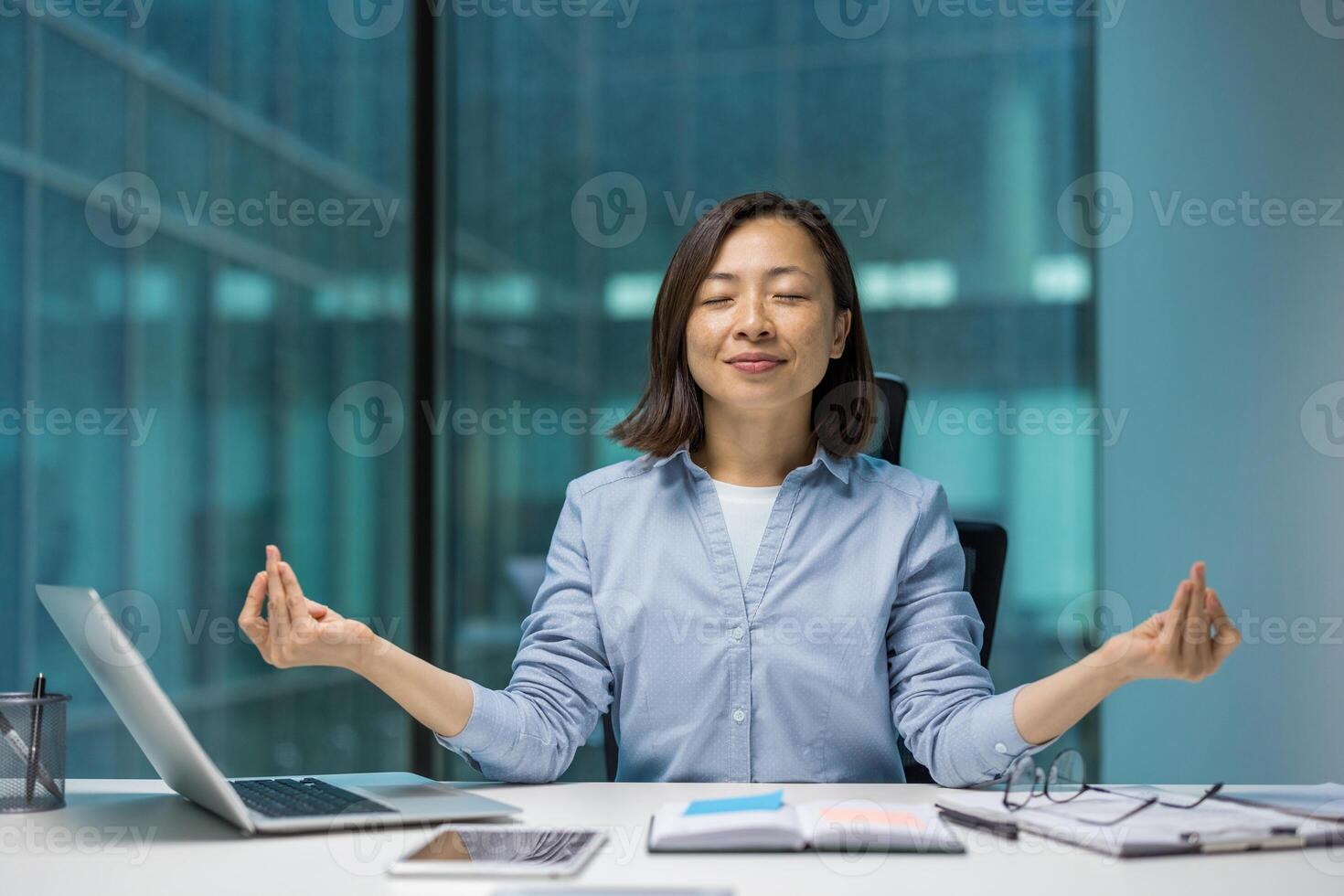 Asian businesswoman practicing meditation at desk for stress relief in office. She seated eyes closed, demonstrating calmness and mindfulness. Laptop and phone on desk create business environment. photo