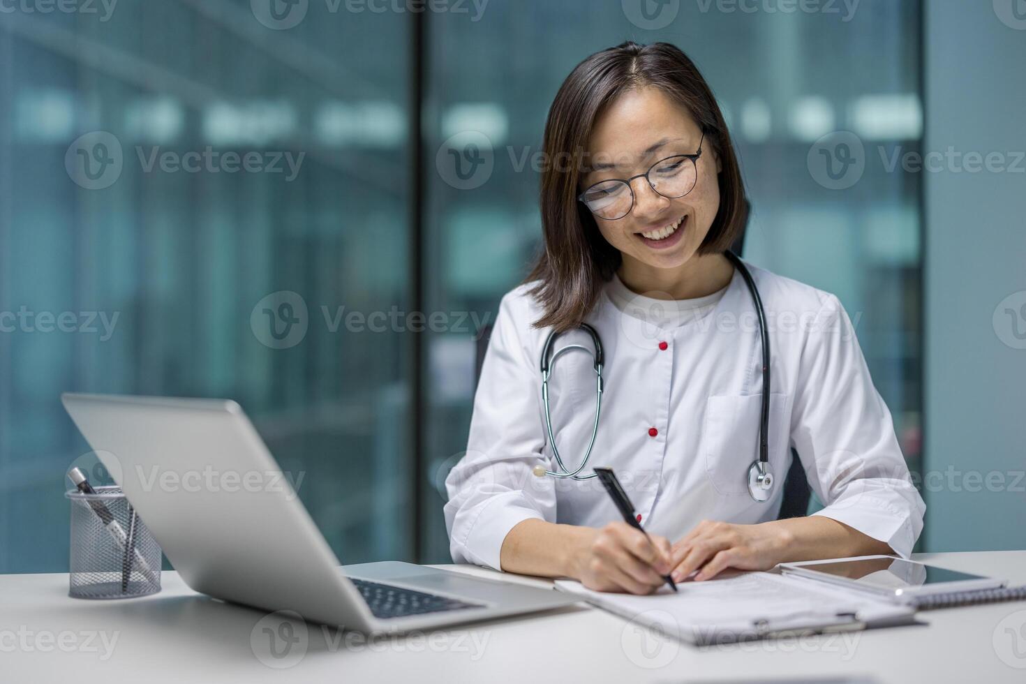 Asian woman doctor with a stethoscope, working on a laptop and taking notes in a professional setting. She appears focused and engaged in her tasks. photo