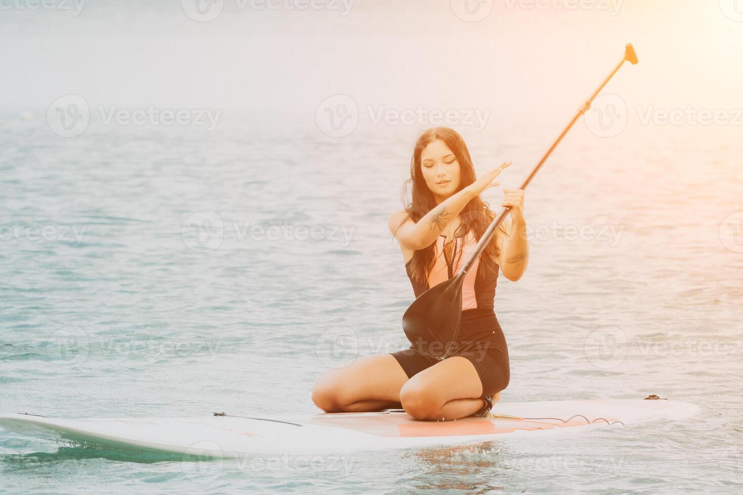 Close up shot of beautiful young caucasian woman with black hair and freckles looking at camera and smiling. Cute woman portrait in a pink bikini posing on sup board in the sea photo