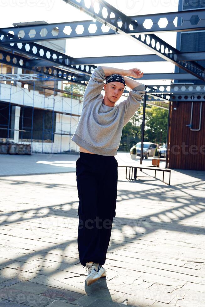 A young man in a grey sweatshirt and black pants stands in an urban setting, his arms crossed behind his head photo