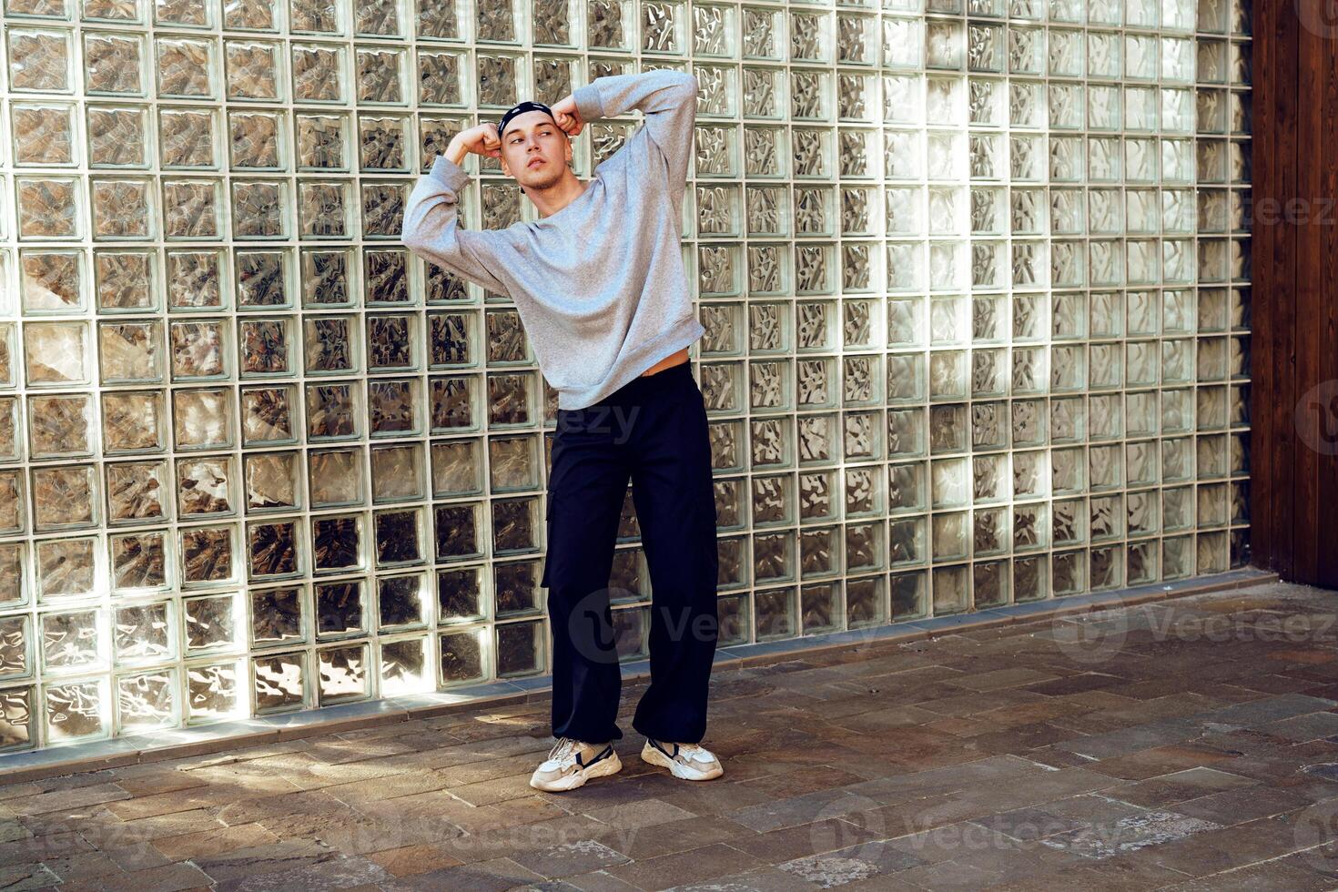 A young man wearing a gray sweater and black pants stands in front of a glass block wall, posing with one arm raised above his head photo
