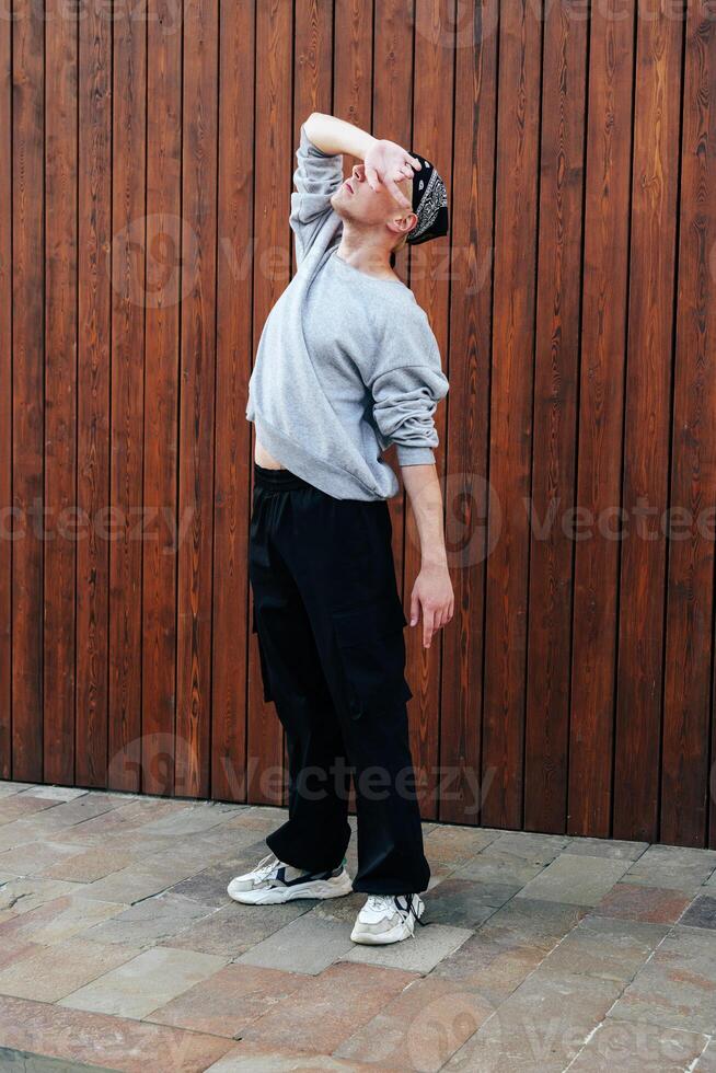 A person in a grey sweatshirt and black pants is striking a pose in front of a wooden wall photo