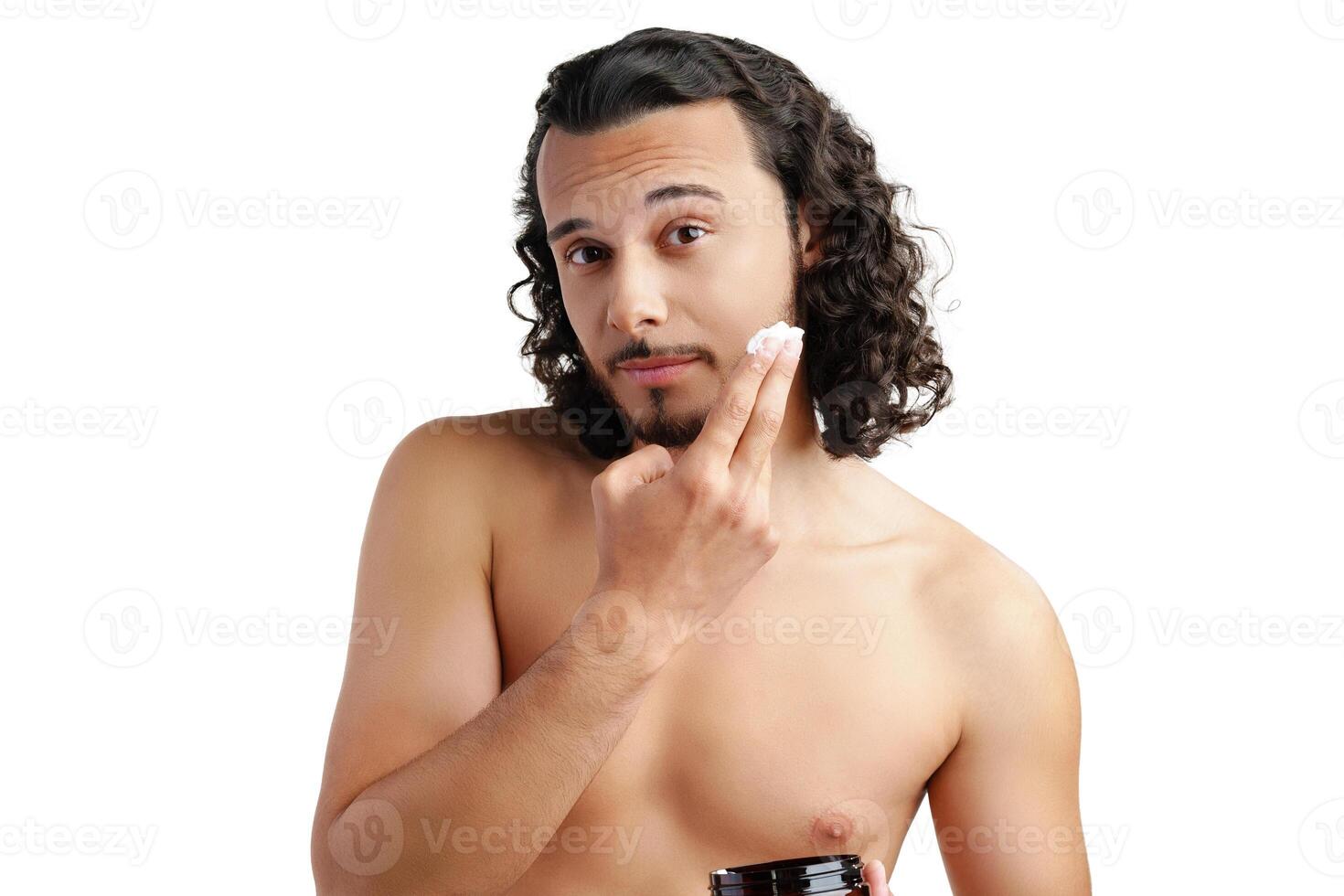 A young man applying skincare cream on his face with a focused expression, demonstrating personal grooming at home photo