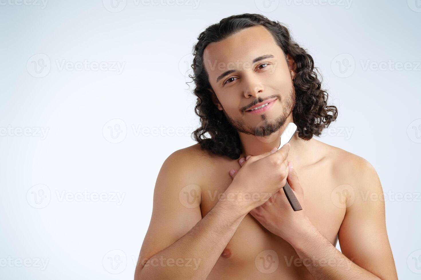 A man with curly hair grooming his beard in a well-lit space while using a straight razor photo
