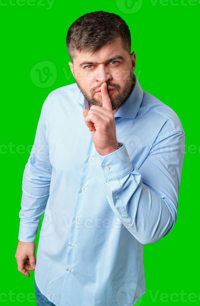 A man in a blue shirt gestures for silence with a finger on his lips against a bright green background photo