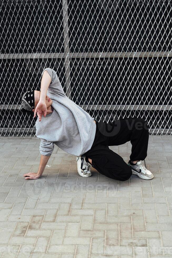 A young man wearing a gray sweatshirt and black pants is performing a dance move in front of a wire fence photo