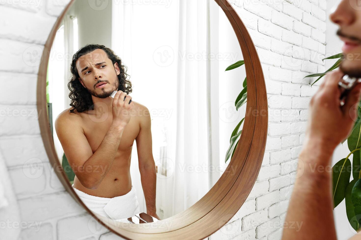 A man with curly hair grooming himself in the bathroom while looking into a round mirror photo