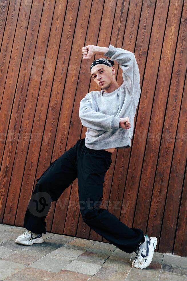 A man in a gray sweatshirt and black pants dances in front of a wooden wall photo